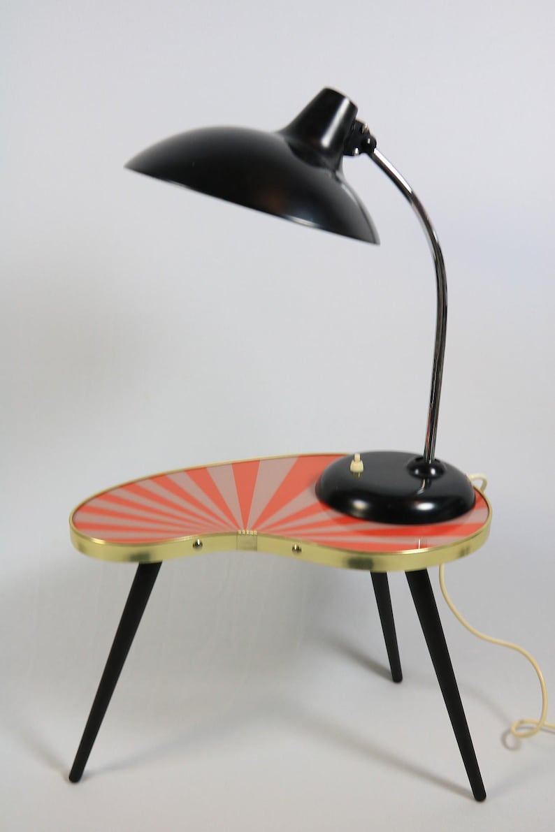 May include: A mid-century modern accent table with a built-in lamp. The table has a kidney-shaped top with a red and pink sunburst design, brass trim, and black legs. The lamp has a black shade and a chrome gooseneck.
