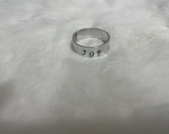 May include: A silver ring with the word "JOY" stamped on the outside. The ring is set against a soft, white background. The ring is a simple band style.