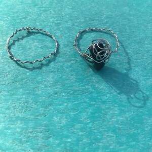 Puede incluir: Dos anillos de alambre plateado, uno con una piedra negra envuelta en alambre. Los anillos están sobre una superficie turquesa.