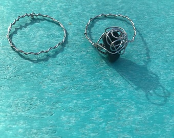 May include: Two silver wire rings, one with a black stone wrapped in wire. The rings are on a turquoise surface.