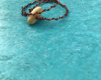 May include: A handmade ring with a light-coloured, oval-shaped stone wrapped in copper wire. The wire is twisted around the stone and forms the ring's band. The ring rests on a turquoise surface.