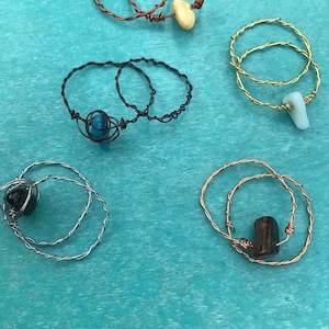 Puede incluir: Cinco anillos envueltos en alambre con piedras de diferentes colores. Los anillos están hechos de alambre de cobre, plata y oro. Las piedras son de color amarillo claro, azul, negro, marrón y verde.