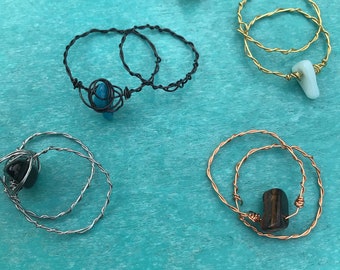 May include: Five wire wrapped rings with different coloured stones. The rings are made with copper, silver, and gold wire. The stones are a light yellow, blue, black, brown, and green.