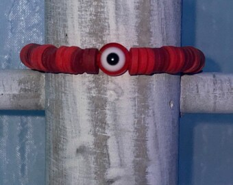 May include: A red and white beaded bracelet with a blue evil eye charm. The bracelet is on a white wooden post.