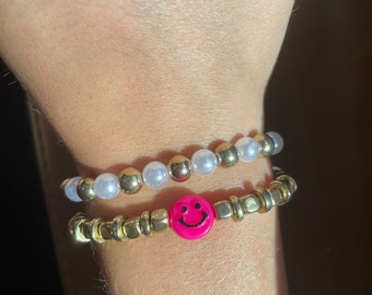 May include: Two bracelets on a wrist. The top bracelet is made of white and gold beads. The bottom bracelet is made of gold beads with a pink smiley face charm.