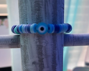 May include: A blue and white beaded bracelet featuring a blue evil eye charm. The bracelet is made from small, round beads in various shades of blue and white.