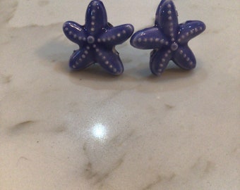 May include: Two purple ceramic starfish with white dots. The starfish are on a white marble surface.