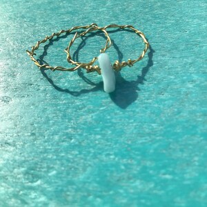 Puede incluir: Dos anillos de alambre dorado envueltos con un dije de piedra azul claro.