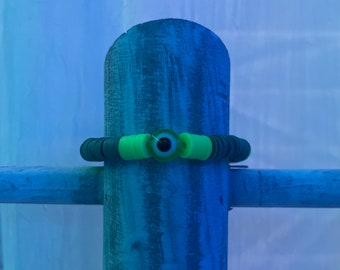 May include: A green and black beaded bracelet featuring an eye-shaped bead at its centre. The bracelet is composed of small, round beads in varying shades of green and black, with a bright green accent. The eye-shaped bead has a black centre and is surrounded by a light green colour.