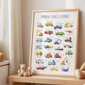 May include: A framed print titled "WORKING VEHICLE ALPHABET" featuring colorful illustrations of various working vehicles, each labeled with a letter of the alphabet. The print is in a light wood frame and is displayed on a wooden table with teddy bears.
