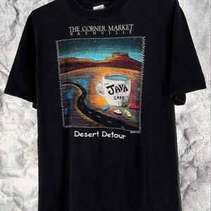 May include: A black short-sleeve t-shirt featuring a graphic print of a desert landscape with a large coffee cup labeled "JAVA CAFE". The text "THE CORNER MARKET NASHVILLE" appears above the graphic, and "Desert Detour" is printed below it.