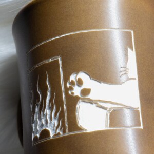 May include: A matte brown ceramic mug featuring a laser-etched design of a cat paw reaching towards a fireplace. The engraving reveals a white base layer beneath the brown surface, creating a high-contrast graphic of flames and a feline limb.