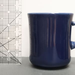 May include: A solid navy blue ceramic mug with a rounded handle, positioned next to a clear plastic ruler that shows the mug stands at a height of approximately 9 cm. The mug has a smooth, glossy finish and a simple, cylindrical shape.