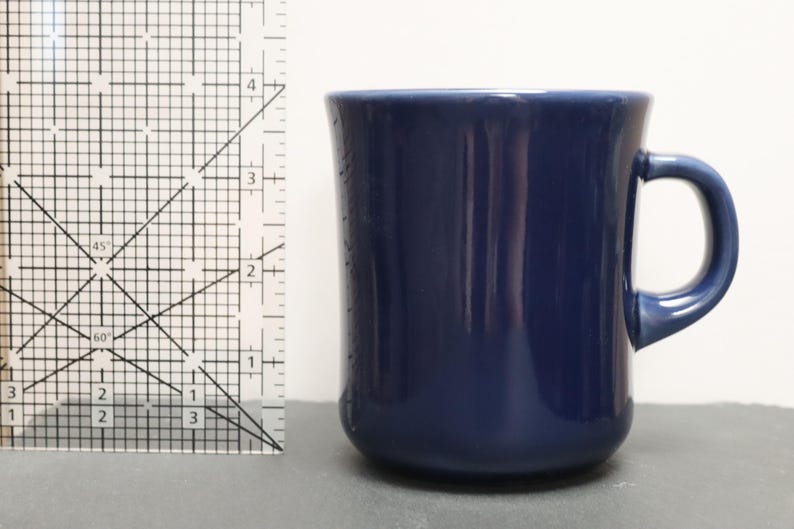 May include: A solid navy blue ceramic mug with a rounded handle, positioned next to a clear plastic ruler that shows the mug stands at a height of approximately 9 cm. The mug has a smooth, glossy finish and a simple, cylindrical shape.