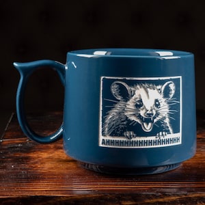 May include: A blue ceramic mug with a raccoon illustration in a white frame. The raccoon is smiling with the word "AHHHHHHHHHHHHH" below it. The mug has a curved handle and sits on a wooden surface.
