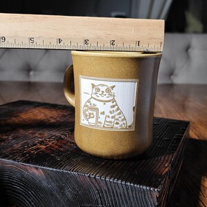 May include: A mustard-coloured ceramic mug featuring a white square graphic of a cat holding a cup. A wooden ruler is positioned above the mug, showing a height of approximately 8.9 cm from the base to the top rim of the ceramic vessel.