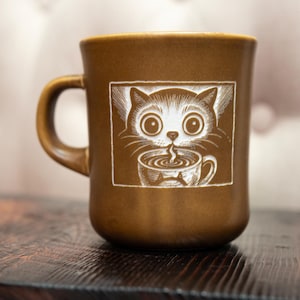 May include: A brown ceramic mug featuring a white graphic illustration of a cat with large eyes holding a small cup of steaming liquid. The mug has a smooth, matte finish and a curved handle on the left side, centred against a neutral background.
