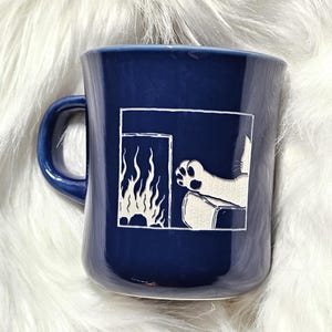 May include: A deep blue ceramic mug featuring a white etched graphic of a cat paw reaching towards a fireplace with visible flames. The design is contained within a square border on the front of the mug, which has a curved handle on the left side.