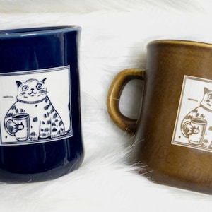 May include: Two ceramic mugs, one dark blue and one brown, each featuring a square graphic of a cat holding a smaller mug. The cat illustration is rendered in a simple line style, and the mugs have a smooth, glossy finish with a curved handle.