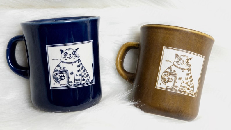 May include: Two ceramic mugs, one dark blue and one brown, each featuring a square graphic of a cat holding a smaller mug. The cat illustration is rendered in a simple line style, and the mugs have a smooth, glossy finish with a curved handle.