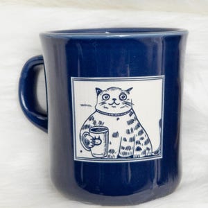 May include: A blue ceramic mug with a white illustration of a cat holding a cup of coffee. The cat is wearing a sweater and has a happy expression.