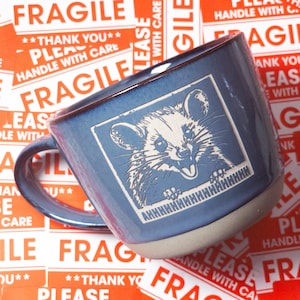 May include: A blue ceramic mug with a white opossum illustration. The mug has a handle and a white rim. The opossum is smiling and has a white face with black eyes and nose. The mug is surrounded by red and white "fragile" stickers.