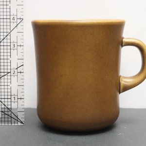 May include: A solid brown ceramic mug with a smooth finish and a curved handle. A clear plastic ruler is positioned to the left of the mug, showing the height of the mug is approximately 10 cm tall from the base to the top rim.
