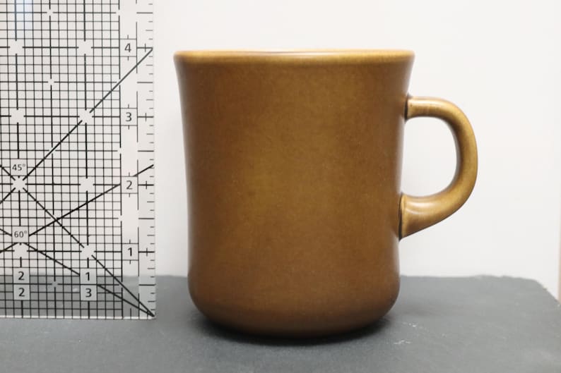 May include: A solid brown ceramic mug with a smooth finish and a curved handle. A clear plastic ruler is positioned to the left of the mug, showing the height of the mug is approximately 10 cm tall from the base to the top rim.