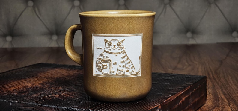 May include: A mustard-brown ceramic mug featuring a white rectangular inset panel. The panel displays a line-art illustration of a seated cat holding a smaller mug. The mug has a smooth, rounded handle and a consistent earthy tone throughout its body.