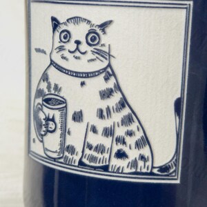 May include: A dark blue ceramic mug featuring a rectangular white panel with a black line drawing of a wide-eyed cat holding a small mug. The cat has a striped pattern on its body and is wearing a collar. The illustration is framed by a thin border.