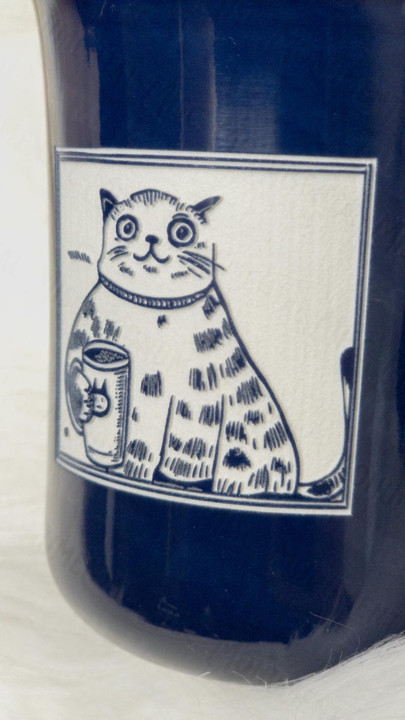 May include: A dark blue ceramic mug featuring a rectangular white panel with a black line drawing of a wide-eyed cat holding a small mug. The cat has a striped pattern on its body and is wearing a collar. The illustration is framed by a thin border.