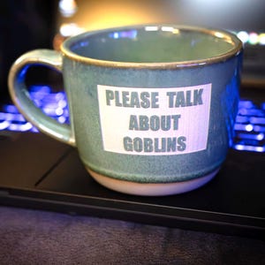 May include: A teal ceramic mug with a white rectangle that reads "PLEASE TALK ABOUT GOBLINS." The mug has a brown rim and handle, and sits on a black laptop. The keyboard is lit with blue lights.