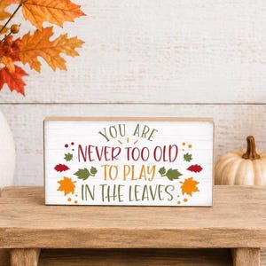 May include: A rectangular wooden sign with a white finish and horizontal wood grain texture. The text reads "YOU ARE NEVER TOO OLD TO PLAY IN THE LEAVES" in green, red, and orange, surrounded by colorful autumn leaf illustrations.
