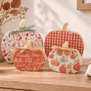 May include: Four decorative wooden pumpkin cutouts. One features a fall leaf pattern with a green ribbon, one has a red gingham pattern, one is orange with text reading Family Time, Hello Fall, Pumpkins, and one displays a small pumpkin print.