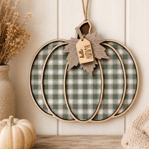 May include: A hanging wooden pumpkin decoration featuring a green and white plaid pattern. The pumpkin has a layered wooden leaf cutout attached to the top, which includes a small rectangular wooden tag with the text hello AUTUMN engraved.