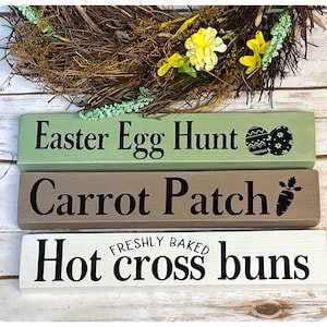 May include: Three wooden signs with Easter-themed text. The top sign is green and says "Easter Egg Hunt" with a black and white egg graphic. The middle sign is brown and says "Carrot Patch" with a black carrot graphic. The bottom sign is white and says "Hot cross buns" with the words "Freshly Baked" in smaller text above.