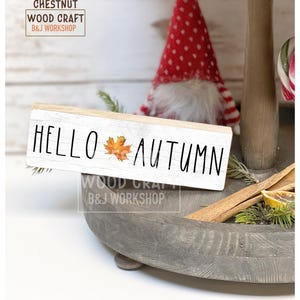 May include: A rectangular white wooden sign with a distressed finish featuring the black text HELLO AUTUMN and a small orange maple leaf graphic in the center. The sign is positioned on a gray tiered tray alongside a red and white gnome.