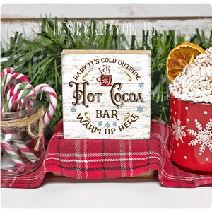 May include: A rustic wooden sign with the text "Hot Cocoa Bar, Warm Up Here" in brown and gold lettering. The sign is on a red plaid cloth, next to a red mug with snowflakes and a jar of candy canes. The sign is a winter-themed decoration.