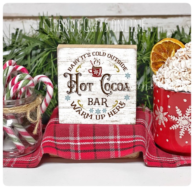 May include: A rustic wooden sign with the text "Hot Cocoa Bar, Warm Up Here" in brown and gold lettering. The sign is on a red plaid cloth, next to a red mug with snowflakes and a jar of candy canes. The sign is a winter-themed decoration.