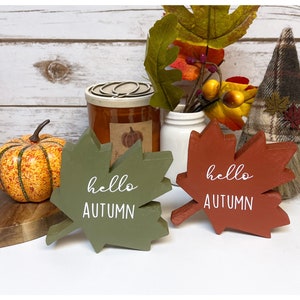 May include: Two wooden leaf-shaped signs with the text "hello autumn" in white paint. The signs are painted in green and red and are sitting on a wooden surface.