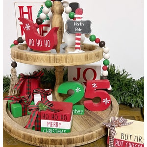 Könnte beinhalten: Ein zweistöckiges Holztablett, das für Weihnachten dekoriert ist. Die obere Ebene zeigt einen Schlitten aus Holz, einen Schneemann und ein Schild mit der Aufschrift "Nordpol". Die untere Ebene zeigt ein Holzschild mit der Aufschrift "JO", eine grüne Holznummer "2", eine rote Holznummer "5", einen Stapel Holzblöcke mit der Aufschrift "HO HO HO FROHE WEIHNACHTEN" und einen Stapel Holzblöcke mit der Aufschrift "BUT FIRST HOT CHOCOLATE".