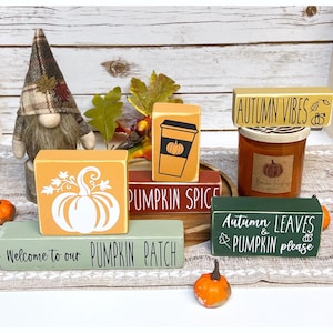 May include: An autumnal display featuring wooden blocks with fall-themed phrases and images, alongside a candle and a gnome ornament. The blocks are painted in shades of orange, green, and brown, creating a seasonal atmosphere.