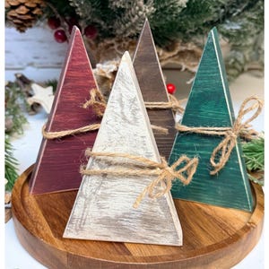 May include: Four wooden Christmas tree decorations in burgundy, white, brown, and green, arranged on a wooden tray. Each tree is tied with twine. The background features pine branches and berries.