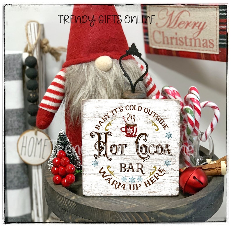 May include: A rustic wooden sign with the text "Hot Cocoa Bar, Warm Up Here" is displayed. The sign is surrounded by Christmas decorations, including a gnome, candy canes, and a small Christmas tree. The sign is on a wooden tray.