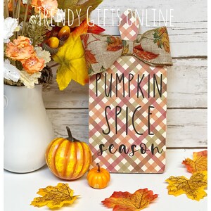 May include: A decorative wooden block with a plaid pattern and the words "PUMPKIN SPICE season". It is topped with a bow featuring fall leaves. Two small pumpkins and fall leaves are in front of the block. A vase of flowers is to the left.