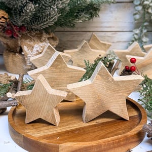 May include: Several wooden star decorations of varying sizes, with a natural wood grain finish. They are arranged on a wooden tray. Greenery and red berries are placed around the stars, suggesting a festive, holiday theme.