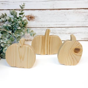 May include: Three natural light-coloured wooden pumpkin cutouts with visible wood grain patterns. The pumpkins are arranged in a group, each featuring a distinct stem and rounded shape, showcasing the raw texture and organic knots of the wood.