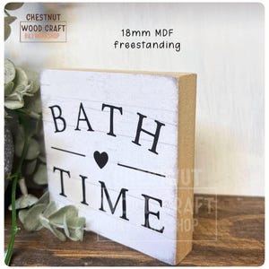 May include: A freestanding white wooden sign with the words "BATH TIME" in black letters, a black heart, and a line separating the words. The sign is 18mm MDF and has a rustic, distressed look.