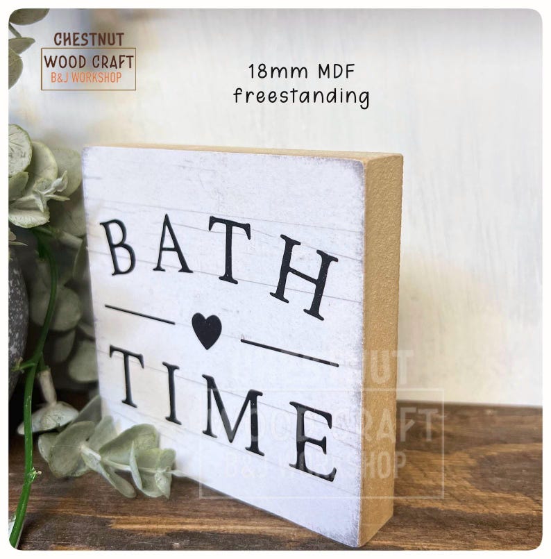 May include: A freestanding white wooden sign with the words "BATH TIME" in black letters, a black heart, and a line separating the words. The sign is 18mm MDF and has a rustic, distressed look.