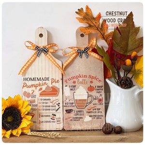 May include: Two wooden recipe boards for pumpkin pie and pumpkin spice latte, each with an orange and black gingham ribbon. The pumpkin pie board lists ingredients like eggs, sugar, and cinnamon. The latte board includes milk, espresso, and pumpkin puree.